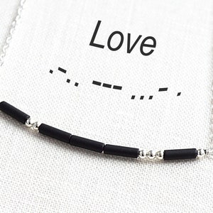 Puede incluir: Un collar de cadena plateada con cuentas negras que deletrean la palabra "Love" en código Morse. El collar está sobre un fondo de tela blanca.