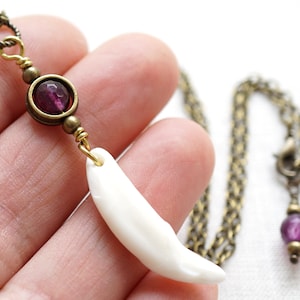 Peut inclure: Un collier avec un pendentif en forme de dent blanche et des accents de pierres précieuses violettes. La chaîne est en laiton et le pendentif est attaché avec un fil de laiton.