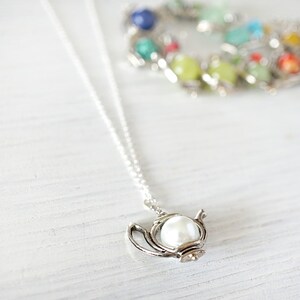 May include: A silver necklace with a teapot pendant featuring a white pearl. The necklace is on a white wooden surface.