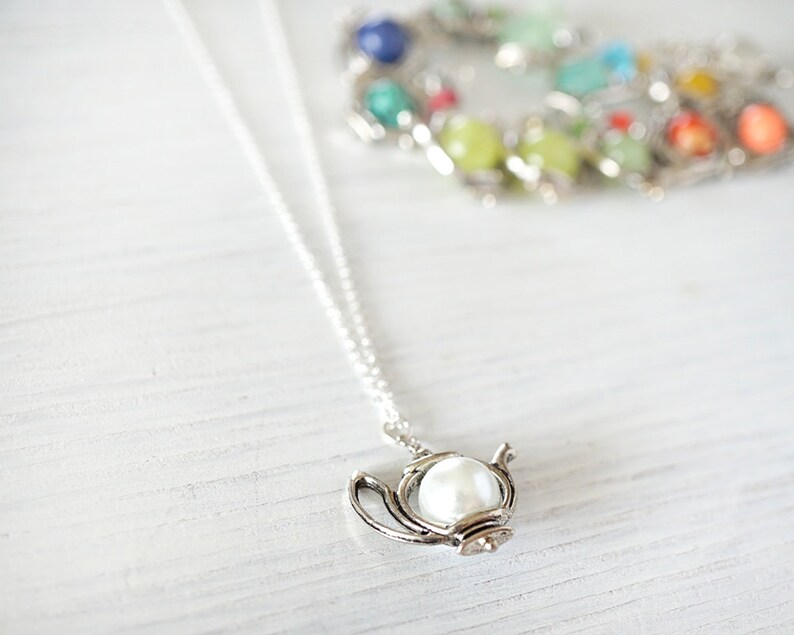 May include: A silver necklace with a teapot pendant featuring a white pearl. The necklace is on a white wooden surface.