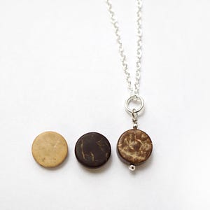 Può includere: Una collana con catena d'argento con un ciondolo rotondo in guscio di noce di cocco marrone. Il ciondolo ha un motivo a venature del legno naturale.