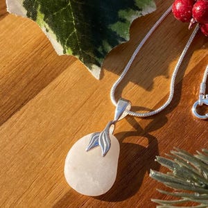 May include: A white stone pendant with a silver leaf design hangs from a silver chain necklace. The pendant is oval shaped and has a smooth, polished surface.