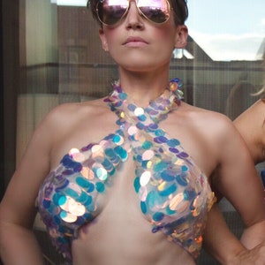 May include: A halter-style crop top constructed from a sheer, transparent base material covered in large, iridescent, circular sequins that reflect blue, purple, and gold tones. The garment features a crisscross neckline design that leaves the midriff exposed.