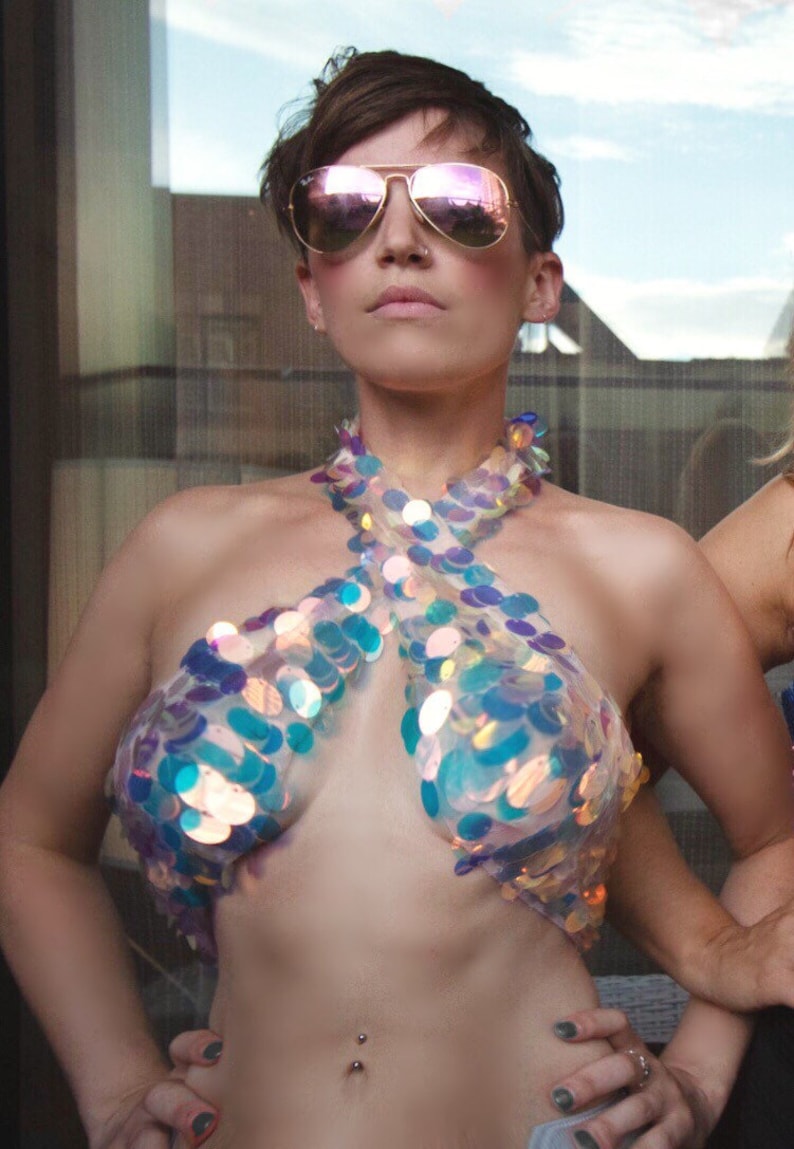 May include: A halter-style crop top constructed from a sheer, transparent base material covered in large, iridescent, circular sequins that reflect blue, purple, and gold tones. The garment features a crisscross neckline design that leaves the midriff exposed.