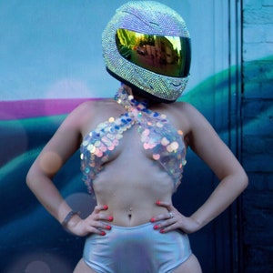 May include: A full-face motorcycle helmet covered in small, reflective silver crystals with a dark, iridescent visor. The outfit includes a halter-style top made of large, translucent, circular sequins and high-waisted, metallic silver shorts.