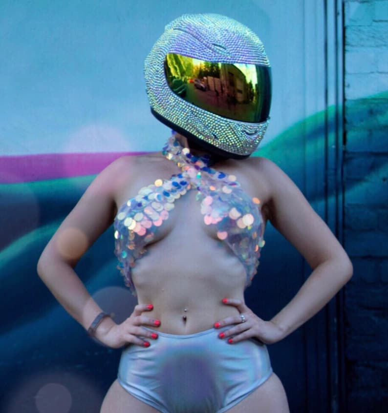 May include: A full-face motorcycle helmet covered in small, reflective silver crystals with a dark, iridescent visor. The outfit includes a halter-style top made of large, translucent, circular sequins and high-waisted, metallic silver shorts.