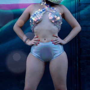 May include: A full-coverage motorcycle helmet covered in shimmering rhinestones with a reflective gold visor, paired with a halter-style top featuring large iridescent sequins and high-waisted silver metallic shorts with a smooth finish.
