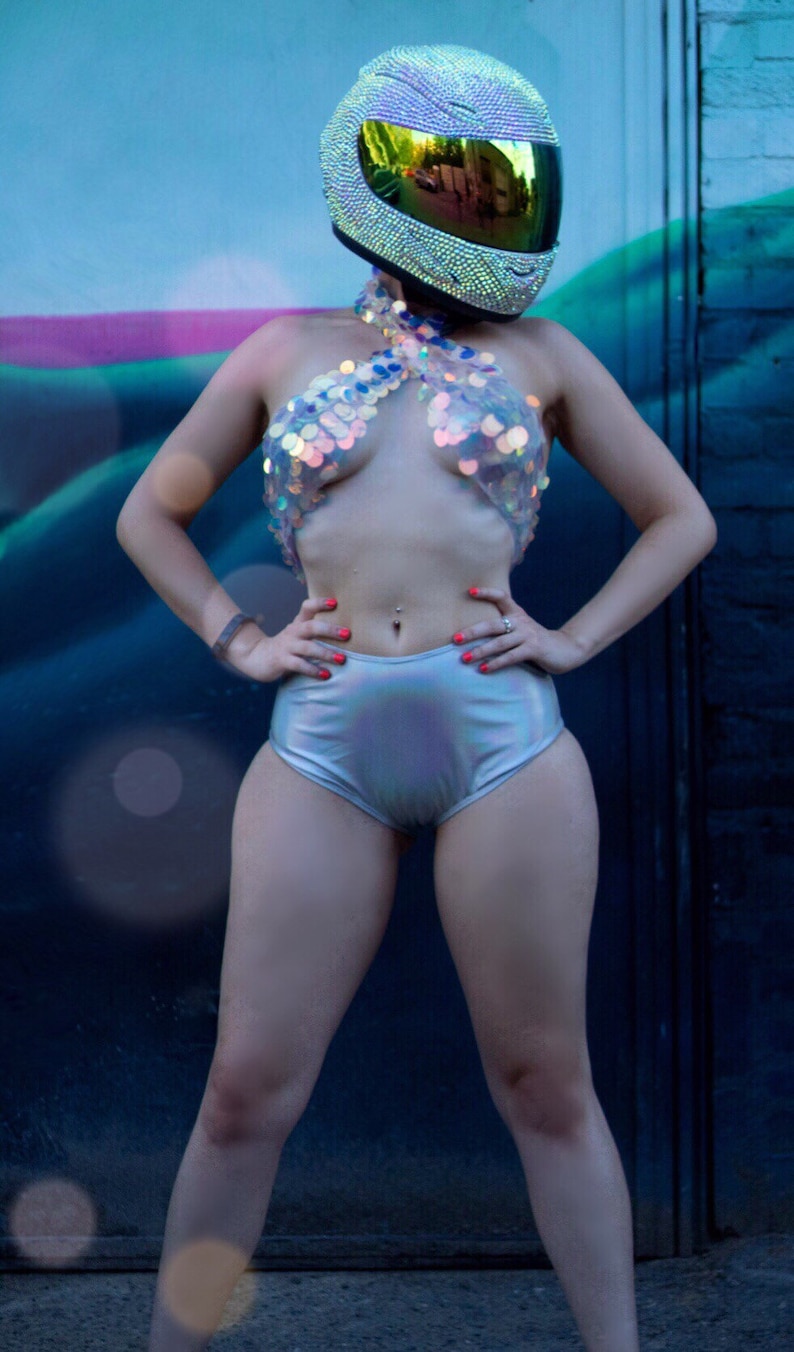 May include: A full-coverage motorcycle helmet covered in shimmering rhinestones with a reflective gold visor, paired with a halter-style top featuring large iridescent sequins and high-waisted silver metallic shorts with a smooth finish.