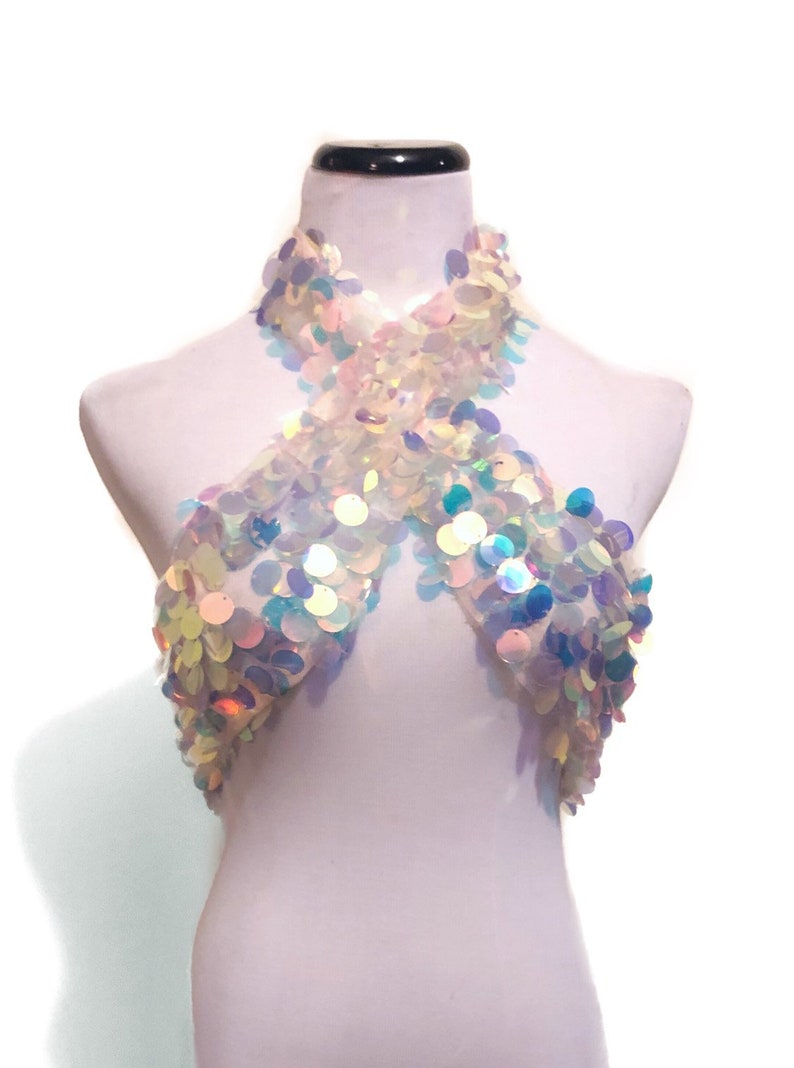 May include: A halter-style crop top covered in iridescent, circular sequins that reflect light in shades of blue, pink, and white. The garment features a crisscross neckline design and is displayed on a white mannequin torso against a plain background.