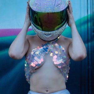 May include: A full-face motorcycle helmet completely covered in small, shimmering iridescent rhinestones with a dark, reflective green visor. The helmet is paired with a halter-style top constructed from large, translucent, circular iridescent sequins.