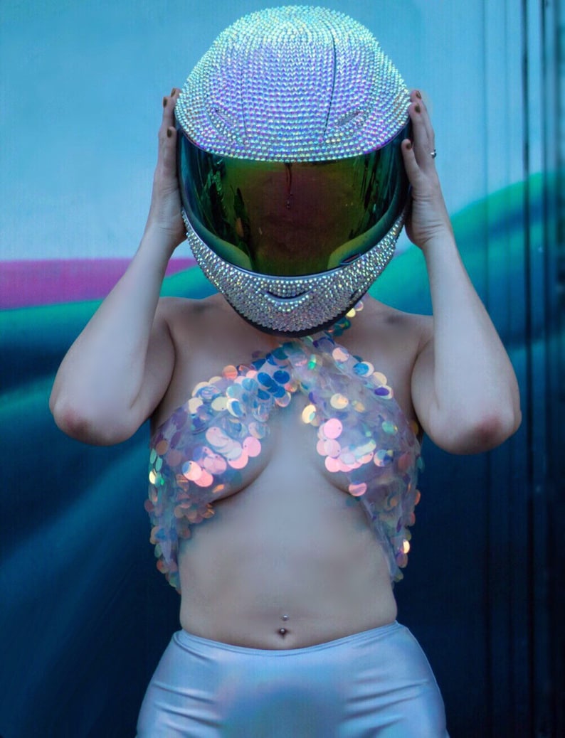 May include: A full-face motorcycle helmet completely covered in small, shimmering iridescent rhinestones with a dark, reflective green visor. The helmet is paired with a halter-style top constructed from large, translucent, circular iridescent sequins.