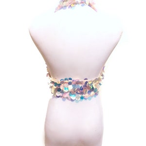 May include: A halter-style crop top covered in iridescent, circular sequins that reflect light in shades of blue, pink, and silver. The garment features a neck strap and a band of overlapping sequins that wrap around the midsection, creating a shimmering, multi-colored effect against a plain white background.