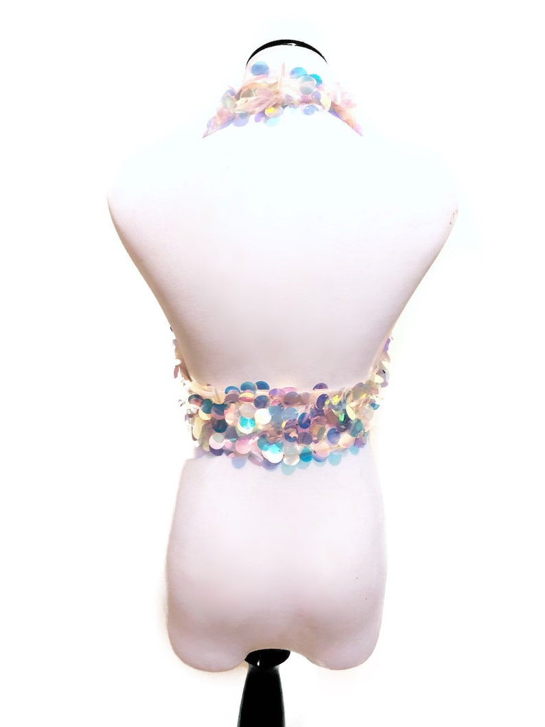 May include: A halter-style crop top covered in iridescent, circular sequins that reflect light in shades of blue, pink, and silver. The garment features a neck strap and a band of overlapping sequins that wrap around the midsection, creating a shimmering, multi-colored effect against a plain white background.