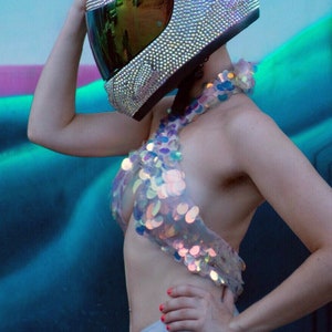 May include: A full-face motorcycle helmet completely covered in small, iridescent, light-reflecting crystals with a dark, reflective visor. The helmet is paired with a halter-style top made of large, circular, iridescent sequins and high-waisted shorts.