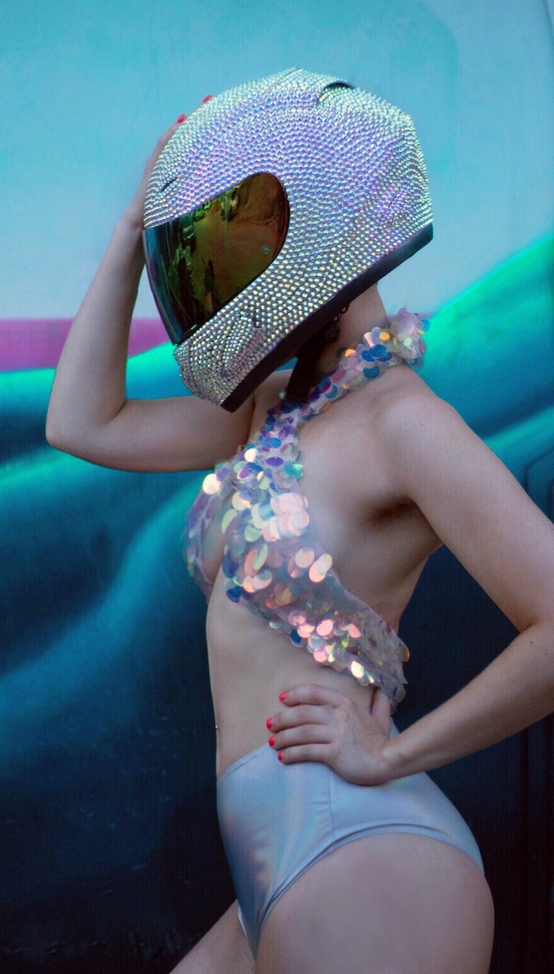 May include: A full-face motorcycle helmet completely covered in small, iridescent, light-reflecting crystals with a dark, reflective visor. The helmet is paired with a halter-style top made of large, circular, iridescent sequins and high-waisted shorts.