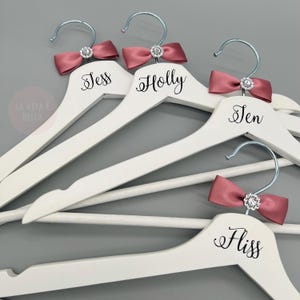 May include: White wooden hangers with silver hooks, each featuring a dusty rose satin bow and a sparkling rhinestone. The names Jess, Holly, Jen, and Fliss are printed on the hangers. The phrase "LA VITA &Egrave; BELLA" is in the corner.
