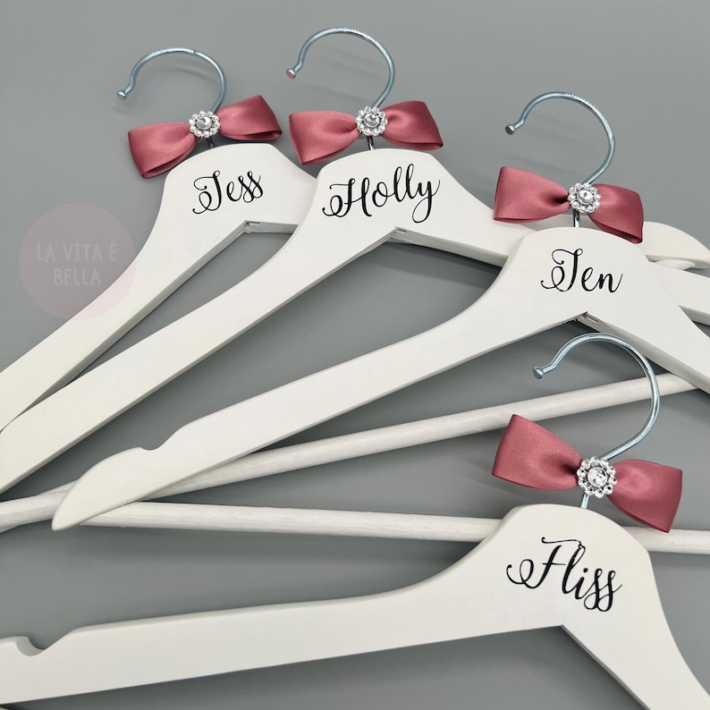 May include: White wooden hangers with silver hooks, each featuring a dusty rose satin bow and a sparkling rhinestone. The names Jess, Holly, Jen, and Fliss are printed on the hangers. The phrase "LA VITA &Egrave; BELLA" is in the corner.