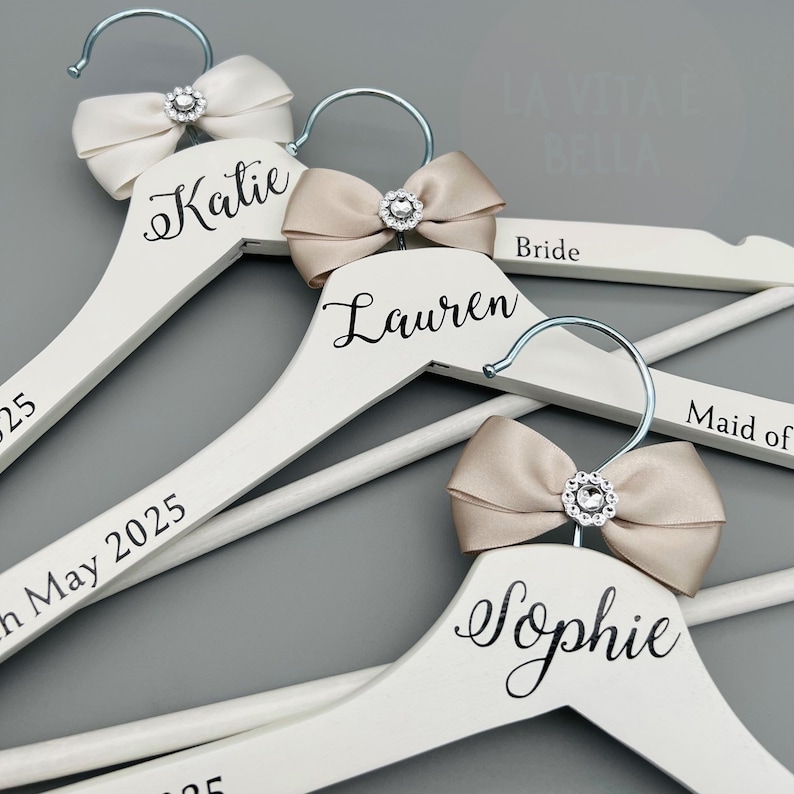 May include: White wooden clothes hangers with silver hooks, decorated with satin bows and rhinestone details. Each hanger is personalised with a name in elegant script, ideal for a wedding. The names Katie, Lauren, and Sophie are visible.