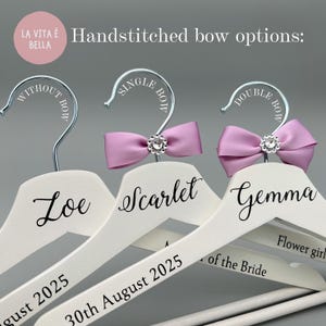 May include: White wooden hangers with silver hooks, personalised with names and dates. Some hangers feature handstitched lavender bows with crystal embellishments. Text includes "Loe", "Scarlet", "Gemma", and "30th August 2025".