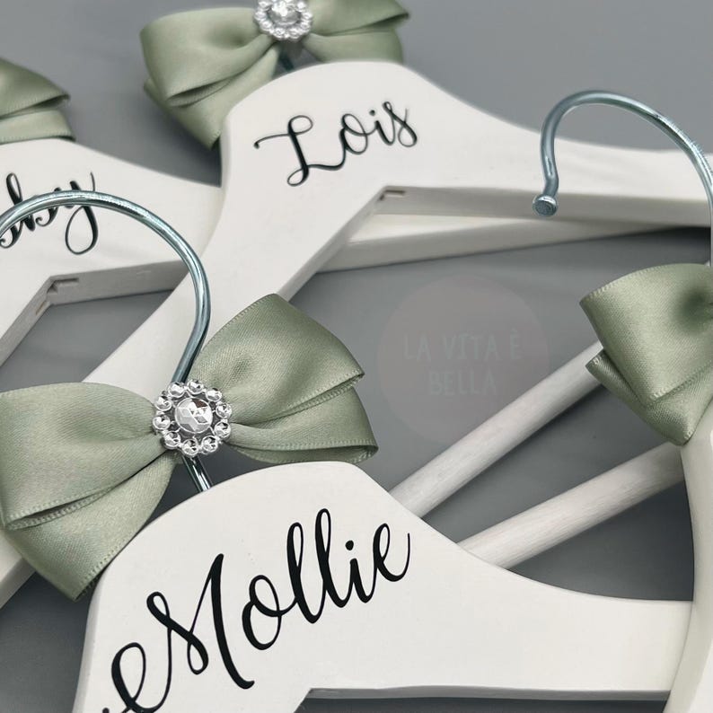 May include: White wooden clothes hangers with silver hooks, decorated with sage green satin bows and sparkling rhinestone details. The names "Lois" and "Mollie" are written in black script on the hangers, ideal for a wedding or special occasion.