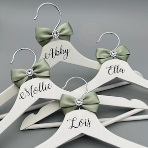 May include: A selection of white wooden coat hangers with silver hooks. Each hanger features a sage green bow with a rhinestone centre. Names are written in black script: Abby, Mollie, Ella, and Lois. The background is a solid grey.