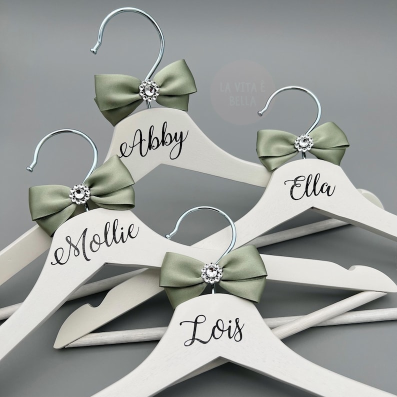 May include: A selection of white wooden coat hangers with silver hooks. Each hanger features a sage green bow with a rhinestone centre. Names are written in black script: Abby, Mollie, Ella, and Lois. The background is a solid grey.