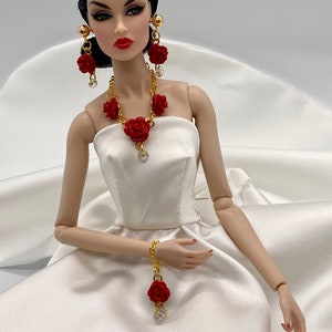 May include: A fashion doll wearing a strapless white satin gown, gold-tone chain necklace with red rose charms and clear beads, matching drop earrings with red roses and gold spheres, and a gold chain bracelet featuring a single red rose charm.