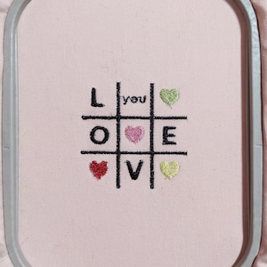 Puede incluir: Diseño bordado en tela rosa en un aro. El diseño es un tablero de tres en raya con la palabra "LOVE" escrita verticalmente. La palabra "you" está escrita sobre la "O". Hay corazones en cada uno de los cuadrados.