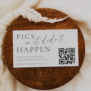 May include: A round, wooden coaster with the text "PICS or it didn't HAPPEN" and a QR code. The coaster is set on a white, textured fabric, with a sprig of dried foliage to the left and a small feather on top.