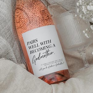 Może przedstawiać: Butelka różowego wina z różową zakrętką i etykietą z napisem "Pairs Well With Becoming a Godmother". Butelka stoi obok przezroczystego kieliszka do wina i bochenka chleba, wszystko na jasnym kocu.