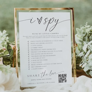 Puede incluir: Una tarjeta de juego de boda enmarcada en oro con el texto "i spy" escrito a mano. La tarjeta incluye una lista de indicaciones fotográficas y un código QR. Descansa sobre una superficie de madera, decorada con vegetación.
