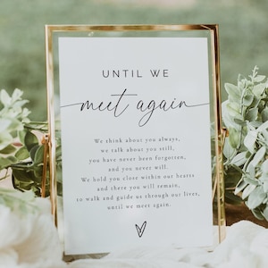 Puede incluir: Un letrero enmarcado con las palabras "UNTIL WE meet again" en escritura elegante. El letrero incluye un mensaje sincero y un símbolo de corazón. El marco es dorado, y el letrero está rodeado de vegetación y tela blanca, sobre una superficie de madera.