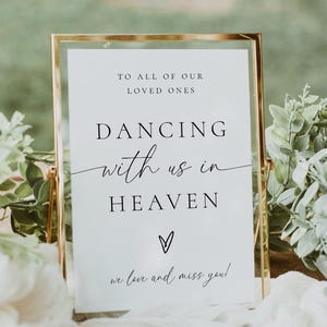 May include: A framed sign with gold trim and the text "DANCING with us in HEAVEN" in black script. The sign is on a wooden surface, surrounded by greenery and white fabric. The sign is for a memorial.