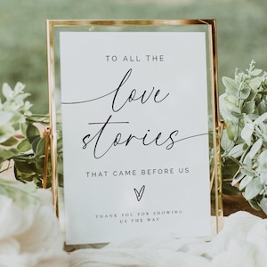 May include: A rectangular sign with gold edging features the words "TO ALL THE love stories THAT CAME BEFORE US". The sign is surrounded by foliage and white fabric, placed on a wooden table, suitable for a wedding or special occasion.