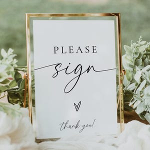 以下が含まれることがあります： 「PLEASE sign」の文字が筆記体で書かれた、金色のフレームのサイン。ハートのシンボルと「thank you！」の文字も。木製のテーブルの上に置かれ、緑と白い布で飾られています。結婚式やイベントに最適です。