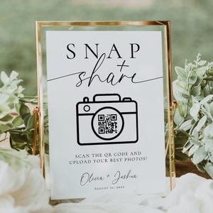 May include: A framed sign with the words "SNAP share" in elegant script and a camera icon with a QR code. The sign also includes the text "SCAN THE QR CODE AND UPLOAD YOUR BEST PHOTOS!" and the names "Olivia + Joshua".