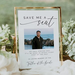 以下が含まれることがあります： 「Save me a seat」の文字と、海辺の男性の写真が入った金色のフレームのサイン。サインには「In loving memory of the Father of the Bride」の文字も含まれています。サインは、緑のある木製の表面に置かれています。