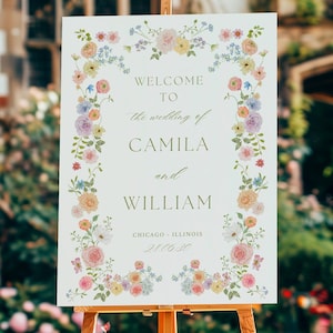 Pode incluir: Um cavalete de madeira exibe uma placa branca com detalhes florais e o texto "Welcome to the wedding of Camila and William, Chicago, Illinois, 2/11/23". A placa é cercada por flores coloridas, com um fundo desfocado de um edifício e uma pessoa.