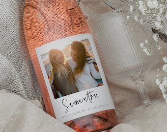Op de afbeelding: Een roze wijnfles met een gepersonaliseerd etiket met een foto van twee vrouwen en de tekst "Samantha, Wil je mijn bruidsmeisje zijn?" De fles ligt op een witte deken met een glas en witte bloemen.