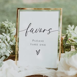 May include: A gold framed sign with the word "favors" in elegant script. Below, it reads "PLEASE TAKE ONE" with a small heart symbol. The sign is on a wooden surface, surrounded by greenery.