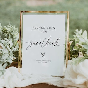 Op de afbeelding: Een goudkleurig bord met de tekst "PLEASE SIGN OUR guestbook" in sierlijke letters. Onder de tekst staat een hartsymbool en de namen "AMELIA + HUDSON" met de datum "08.13.2025". Het bord staat op een houten oppervlak met groen.