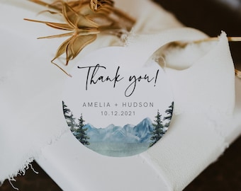 May include: A round white thank you tag with a watercolor illustration of a mountain range and pine trees. The tag says "Thank you! Amelia + Hudson 10.12.2021"
