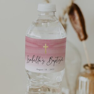 May include: A clear plastic water bottle with a pink and gold label that says "Isabella's Baptism" and "August 10, 2022". The label has a gold cross on it.