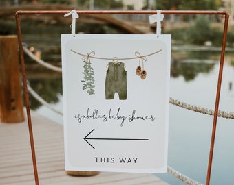 May include: A white sign with a green baby onesie, brown shoes, and green leaves hanging on a clothesline. The sign says "isabella's baby shower" and has an arrow pointing left with the text "THIS WAY".
