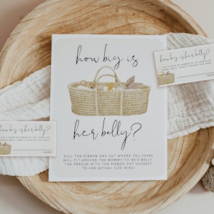 Puede incluir: Una tarjeta blanca con una ilustración de acuarela de una cesta llena de artículos para bebés. La tarjeta dice "How big is her belly?" y tiene instrucciones para un juego de baby shower. La tarjeta está sobre un plato de madera con un paño blanco y una concha.