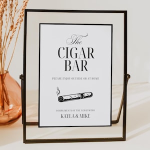 May include: A black framed sign with the text "The Cigar Bar" and a cigar illustration. The sign also includes the text "Please enjoy outside or at home" and "Compliments of the newlyweds Kayla & Mike". A vase with dried plants is in the background.