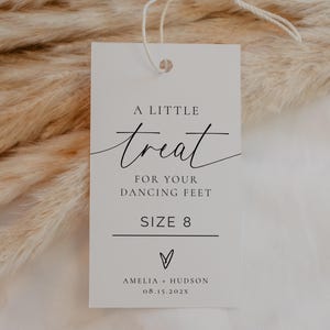 May include: A white rectangular tag with the text "A little treat for your dancing feet" in elegant script. Below, it reads "SIZE 8" and includes a heart symbol and the names "Amelia + Hudson" with a date. The tag is attached to a string.