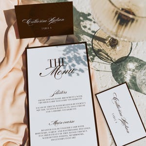 May include: Elegant wedding stationery featuring a menu and place cards. The menu card has a dark border and lists starters, main courses, and dessert options. Place cards display the names "Catherine Hudson" and "Amelia & Hudson".