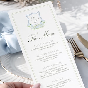 May include: A rectangular white menu card with a gold border, featuring a blue crest with the initials LF and floral accents. The text reads The Menu, First Course, Second Course, Main Course, Dessert, and the names Lauren & Thomas at the bottom.