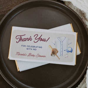 May include: A chocolate bar with a thank you message for celebrating a baby shower. The wrapper features a baseball jersey, bat, glove, and hat. The text reads "Thank You! For celebrating with me! Micaela's Baby Shower."